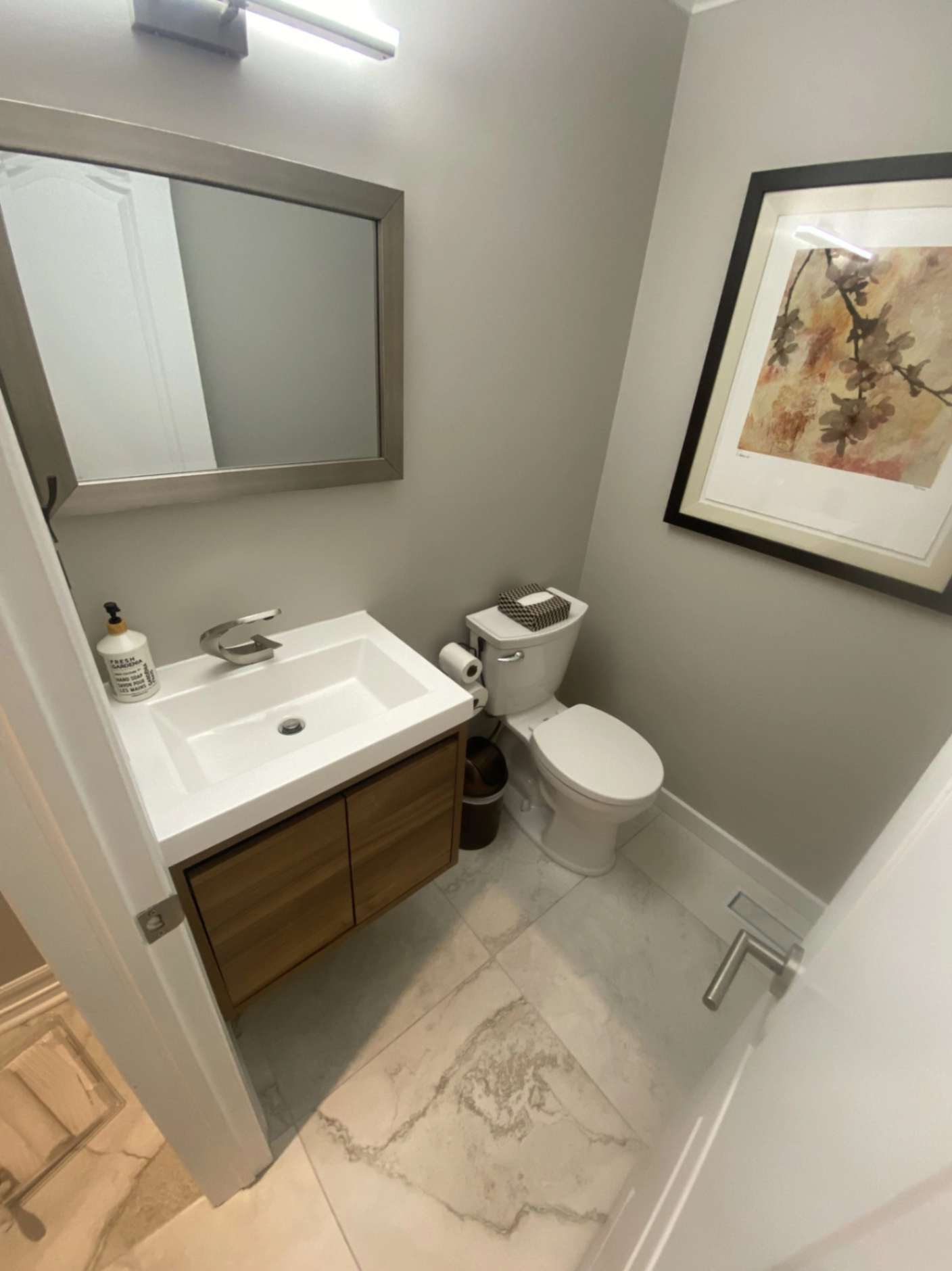 Small modern bathroom with floating vanity, white sink, toilet, wall mirror, and neutral gray walls with framed wall art
